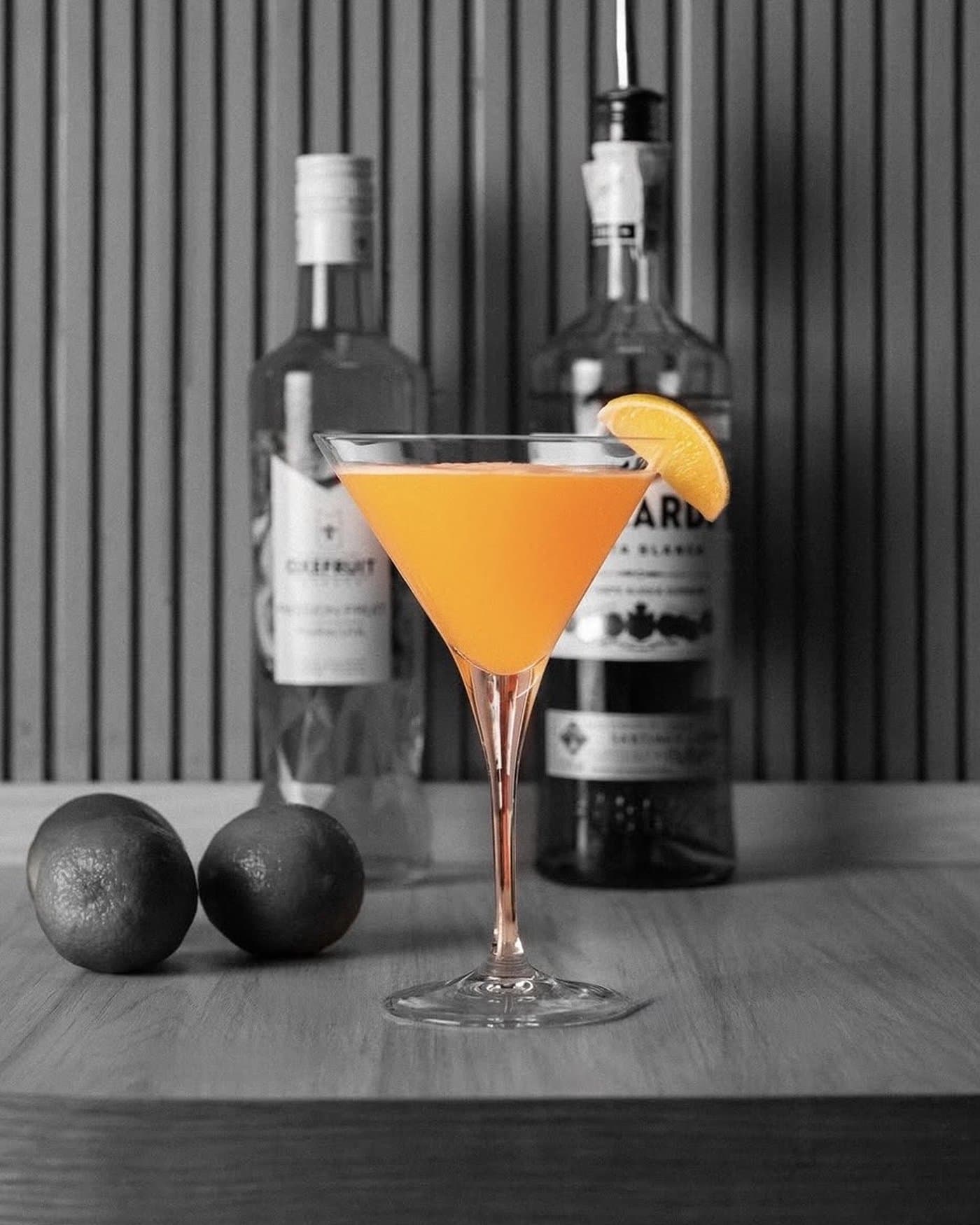 Cóctel de pomelo en copa de martini, con las botellas de la receta detrás en blanco y negro